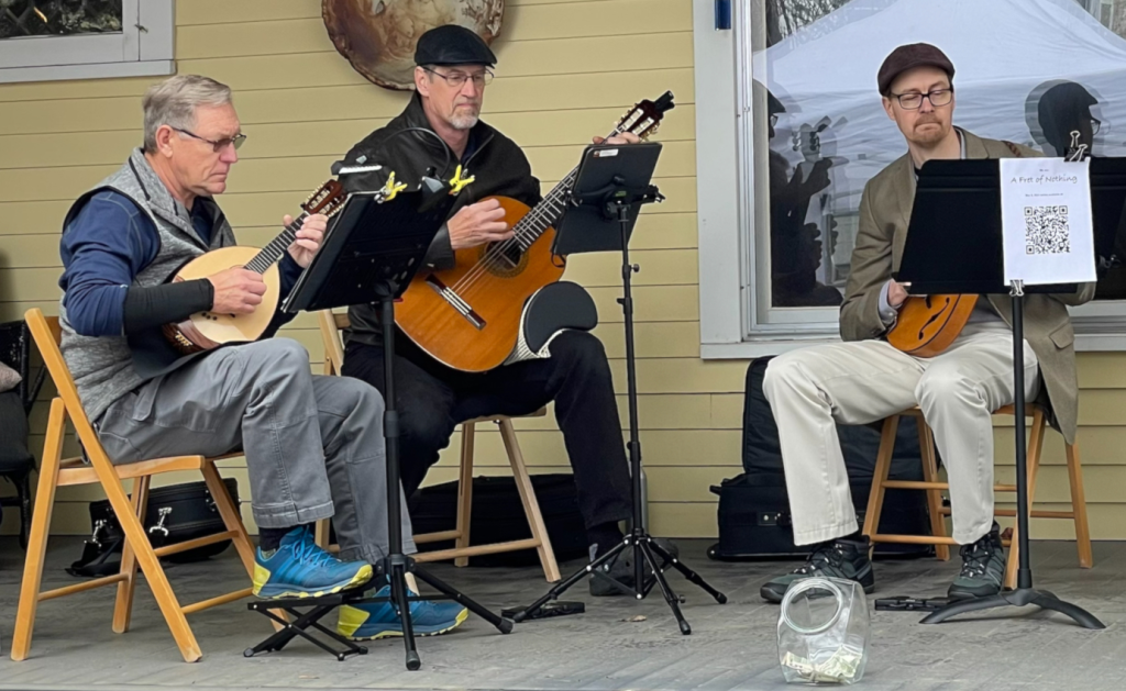 three men sitting on chairs on the porch and playing mandolin, guitar, and mandola